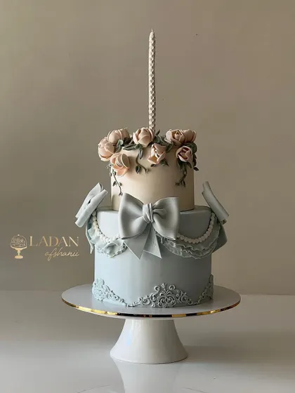 An elegant two-tiered cake featuring soft blue fondant, delicate sugar flowers, and a beautifully crafted bow. This design combines classic piping, molding, and floral techniques.