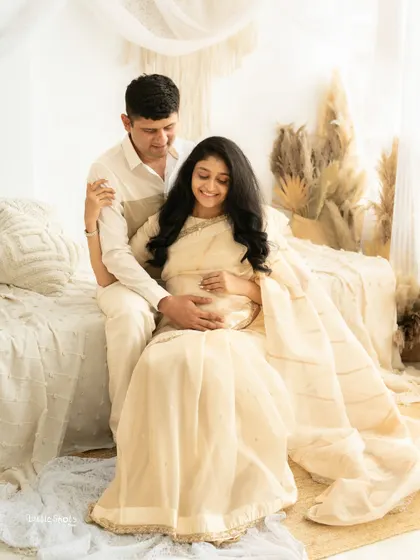 A serene portrait of a couple in a light and airy boho setting. The partner sits behind the mom-to-be, who is wearing a cream saree, as they both gently hold her baby bump.