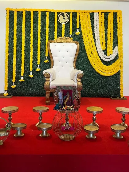 A simple yet elegant Seemantham setup. The decor includes a greenery wall with hanging marigold garlands, a white throne, and a neat arrangement of offering tables and props.