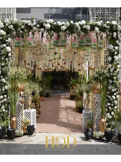 A breathtaking entrance for a Pichwai-themed wedding. The design combines intricate latticework, a ceiling of hanging florals and chandeliers, and a lush green walkway flanked by traditional elements.