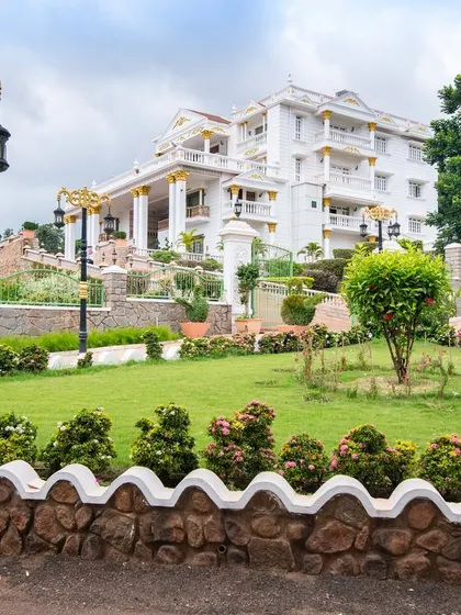 A daytime view of the manicured lawns and gardens of 'The Palace Promised'. The landscape uses a mix of flowering shrubs and formal hedges to create a classic, regal atmosphere.