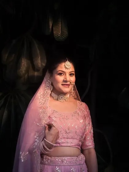 A beautiful portrait of the bride, Vasudha, in her soft pink lehenga, captured with dramatic lighting against a dark, leafy background.
