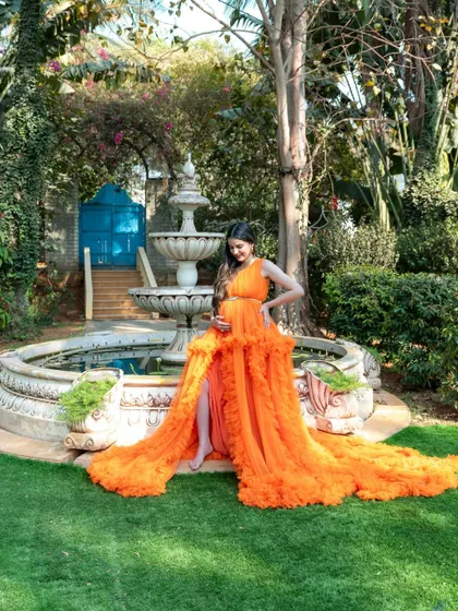 A wide shot of a mom-to-be in a vibrant orange gown posing by a garden fountain.