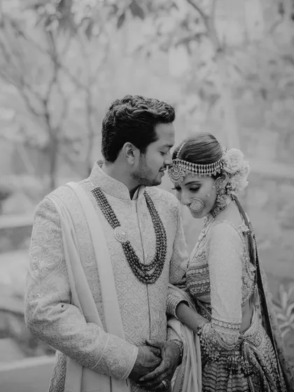 A quiet, intimate black and white portrait of a couple in elegant wedding attire. The groom looks lovingly at the bride, highlighting the fine details of her jewelry and their gentle connection.