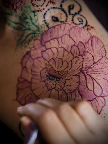 A close up of a large peony flower drawn with colored henna on the shoulder. The fine lines and delicate shading showcase the artistic possibilities of this medium for detailed body art.