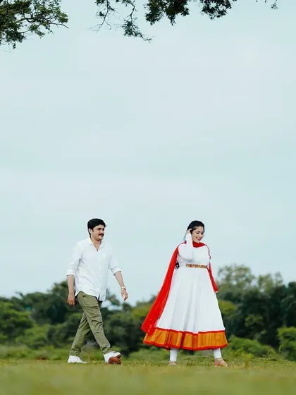 A wide shot of the couple walking in a field, giving a sense of scale and freedom. The vibrant red dupatta adds a beautiful pop of color to the natural landscape.