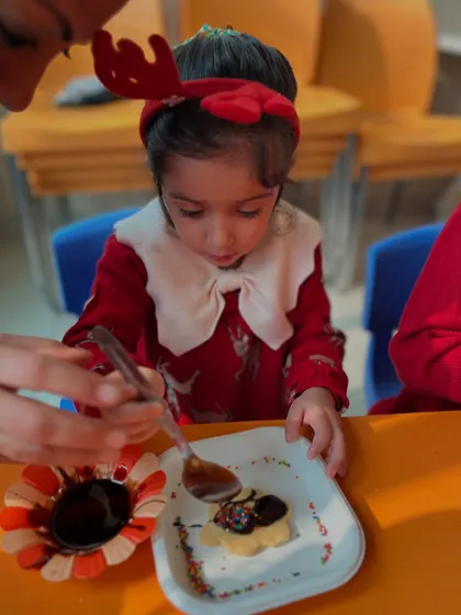 An instructor guides a child's hand as she adds chocolate to her cookie.