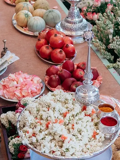 A beautiful display of offerings for a traditional South Indian ceremony. The arrangement includes fresh fruits, flowers, sweets, and other symbolic items, set against a backdrop of lush pink and white florals.