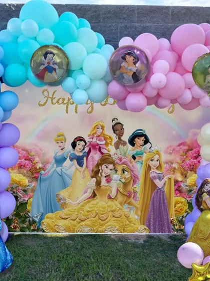 A full and vibrant Disney Princess backdrop for an outdoor party. The setup features a large character poster and a multi-colored balloon arch.
