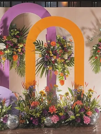 A vibrant and colorful stage setup with orange and purple arches. The tropical flower arrangements and disco balls add a fun and festive energy, perfect for a sangeet or reception party.