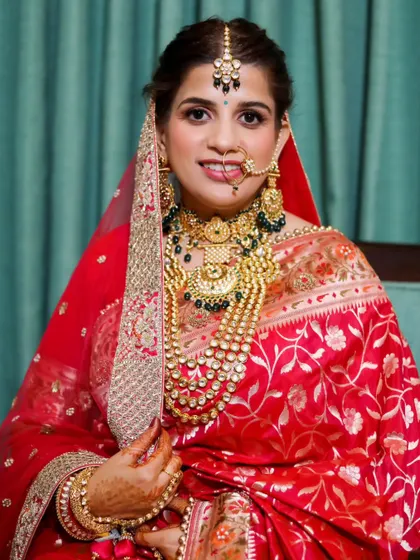 A close-up portrait of a bride in a stunning red Banarasi saree. The focus is on her magnificent gold jewelry and her warm, happy smile.