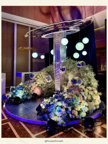 A sculptural centerpiece for the galaxy-themed Sangeet, combining a crystal fringe column with suspended orbs and a base of blue and white florals, creating a futuristic art piece.