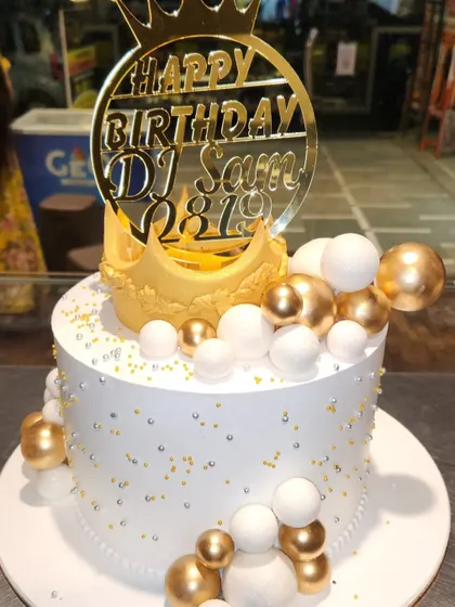 A royal-themed birthday cake perfect for a DJ. This cake features a gold crown, a cascade of white and gold spheres, and a custom acrylic topper.