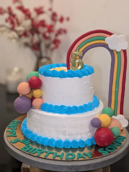 A colorful two-tier rainbow themed cake for a 5th birthday. It's decorated with bright buttercream piping and a large fondant rainbow topper.