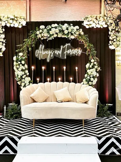 A chic and classic couple seating area for a glam Sangeet. The "Always and Forever" neon sign, circular floral arrangement, and bold black-and-white chevron stage create a modern and stylish focal point.