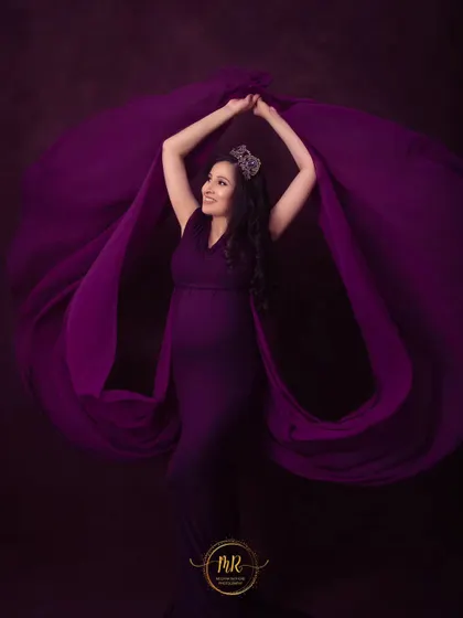 A powerful pose with flowing purple fabric. The mother-to-be wears a crown and holds the fabric aloft, creating a dynamic and regal maternity portrait.
