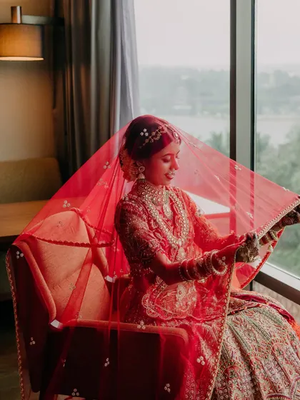 A serene portrait of the bride, Maheswari, seated by a window, her red veil creating a soft, ethereal glow around her.