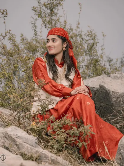A serene portrait of the woman in her red and cream traditional attire, seated among rocks and wild plants, looking thoughtfully into the distance.