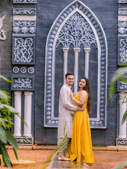 A stylish portrait in front of a unique, gothic-style architectural feature. The couple's bright yellow outfits create a striking contrast with the dark background.