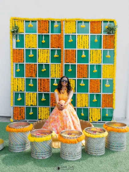 A full view of the bride and her vibrant Haldi setup. The colorful marigold wall and decorative pots create a festive and beautifully coordinated scene.