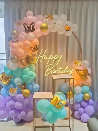 A pastel rainbow butterfly-themed birthday party. This decor includes a circular arch, a 'Happy Birthday' neon sign, and elegant plinth stands.