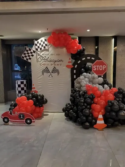 A "Two Fast" car racing theme for a second birthday. The setup includes a custom backdrop with checkered flags, a balloon arrangement in red, black, and grey, and a cutout of a race car.