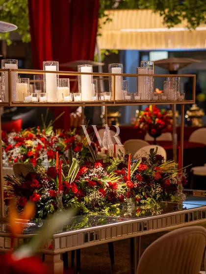 A mirrored dining table reflecting a stunning centerpiece. A long runner of red florals is topped with a gold stand holding dozens of white pillar candles, creating a dramatic and romantic focal point.