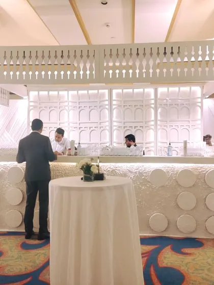 This custom-designed white bar counter features intricate latticework and is flanked by large white floral arrangements, serving as a stylish and functional hub for the reception.