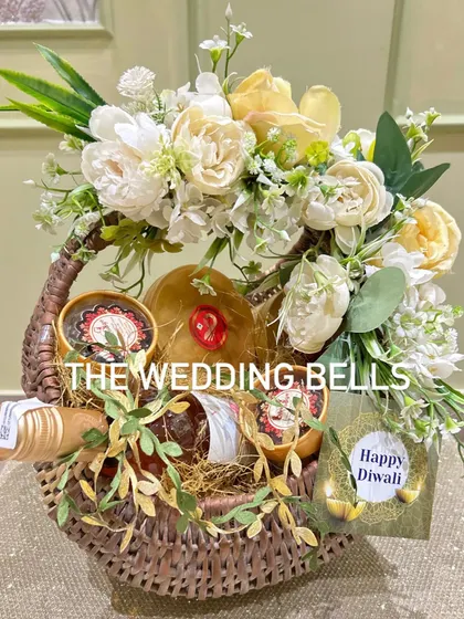 A beautiful and rustic Diwali hamper in a wicker basket. It includes a bottle of wine and other goodies, adorned with a white and yellow floral arrangement.