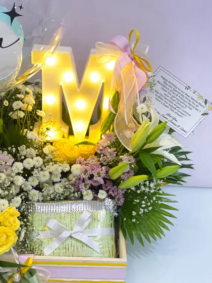 A close-up of the personalized birthday hamper for Meena, showing the glowing 'M' initial nestled among fresh lilies and yellow roses, along with a custom gift card.