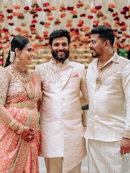 A family photo featuring the couple and their brother. The men's coordinated cream outfits provide a perfect backdrop for the lady's stunning peach and silver saree.