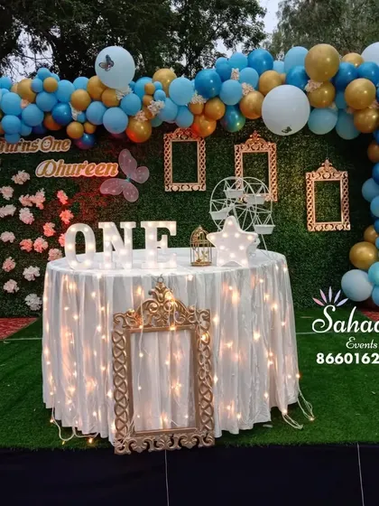 A charming butterfly-themed first birthday party. The decor features a balloon arch in blue and gold, a green hedge backdrop, and delicate butterfly accents for a sweet and whimsical feel.