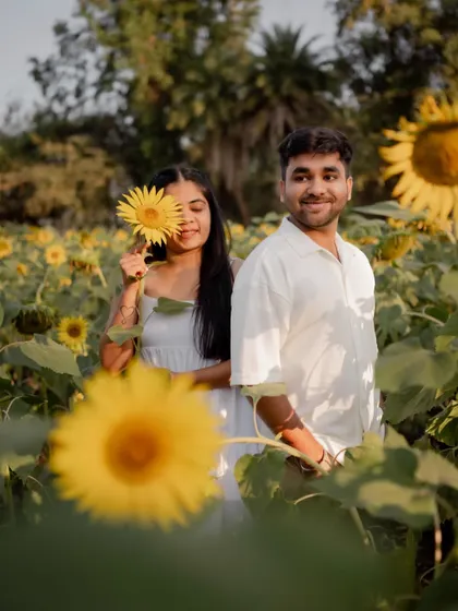 A playful pre-wedding portrait in a sunflower field, with the bride holding a sunflower over her eye while the groom stands beside her.