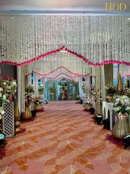 A beautiful walkway for a beautiful beginning. I created this grand entrance with a dense canopy of white and pink floral strings, leading guests down a patterned carpet flanked by traditional brass decor for an elegant wedding reception.
