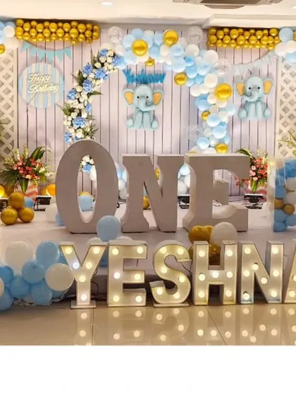A grand first birthday celebration for Yeshnav. The stage is decorated with a massive balloon backdrop in blue, white, and gold, with large light-up letters spelling out the name for a truly show-stopping effect.