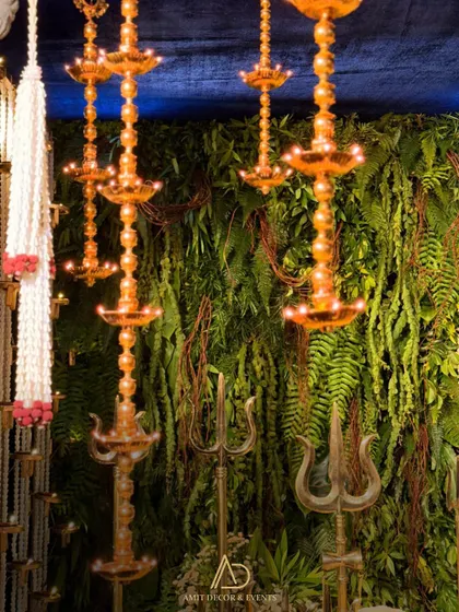 Hanging brass diyas and trishuls are set against a lush green wall, blending traditional divine symbols with natural elements.