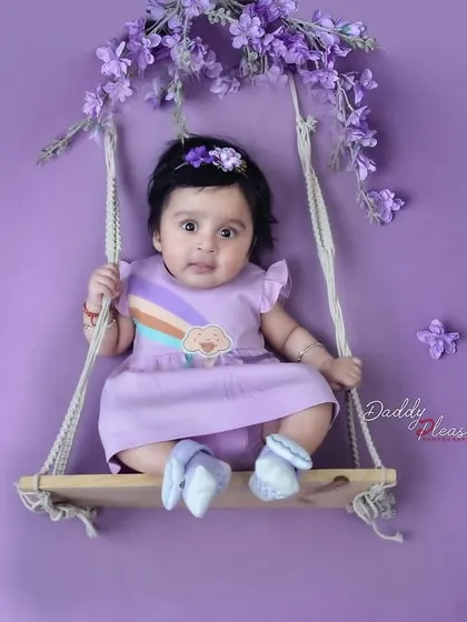 A sweet baby girl in a purple dress sits on a small wooden swing decorated with lavender flowers, all against a matching purple backdrop. This creates a simple yet beautiful color coordinated photo.