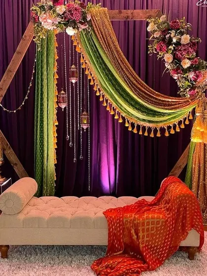 This sangeet or mehendi seating features a modern hexagonal wooden frame. I draped it with colorful green and orange fabrics and accented it with floral bouquets and hanging lanterns for a chic, bohemian vibe.