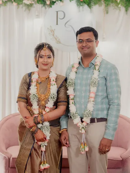 The couple poses for a photo after their engagement ceremony, standing before a simple and elegant backdrop with their initials.