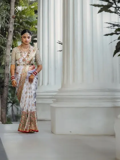 A full-length portrait of the bride walking through a corridor of white pillars. The grand, classical architecture provides a stunning backdrop for her elegant saree.