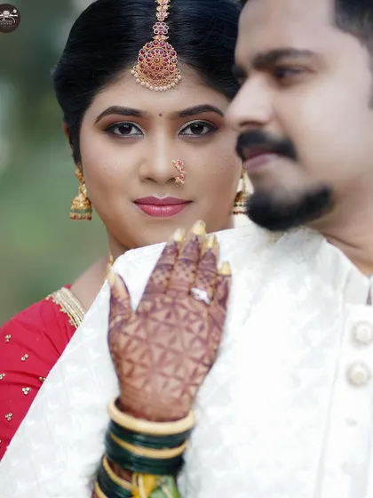 A close-up portrait focusing on the bride looking towards the camera, her hand resting on the groom's shoulder. The shot beautifully displays her bridal makeup, jewelry, and detailed henna design.
