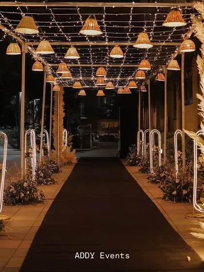 This illuminated passage, featuring a canopy of fairy lights and hanging wicker lamp shades, creates a magical tunnel effect. The black carpet runner and pampas grass arrangements along the sides guide guests through an enchanting experience.