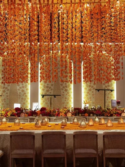 A long dining table set for a traditional feast, with a canopy of hanging marigold strings creating a vibrant and festive ceiling.