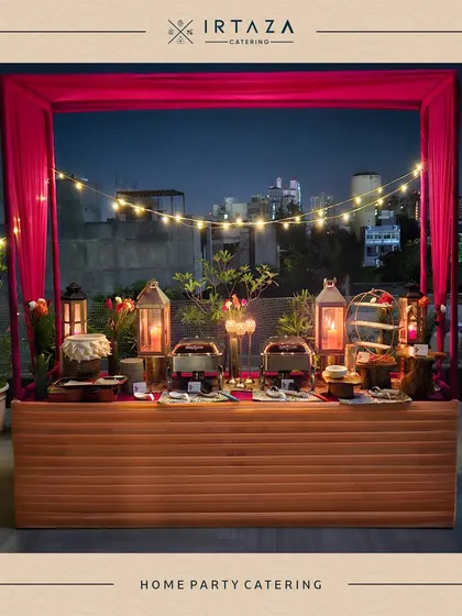 A stunning rooftop home party catering setup. The food station is framed by vibrant pink curtains and string lights, with the city skyline in the background, creating an unforgettable urban party vibe.