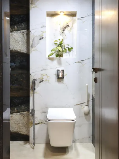 A clean and modern toilet design with a wall-mounted commode. The wall features a combination of white marble and a darker, textured stone, with a lit niche for a plant adding a touch of nature.