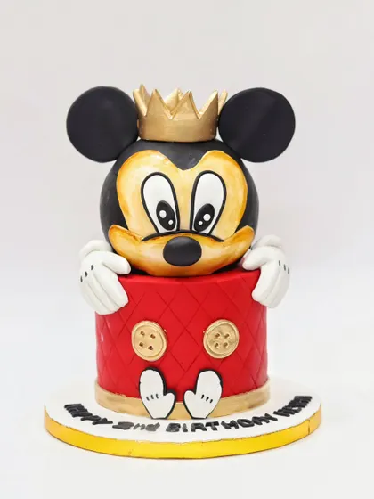 A classic Mickey Mouse cake with a regal twist. This sculpted cake features Mickey's head wearing a crown, with his hands and feet peeking out from the quilted red base.