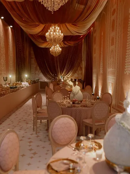 A view of the dining hall, where the ceiling is draped in soft, flowing fabrics and lit by crystal chandeliers. The walls are adorned with custom panels featuring traditional Indian motifs.