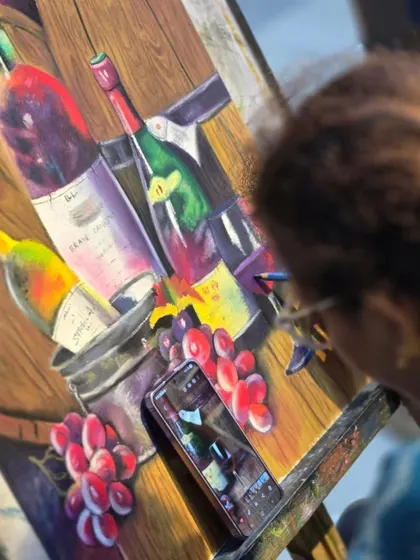 A student works on a colorful still life of wine bottles and grapes, using a reference photo on her phone.