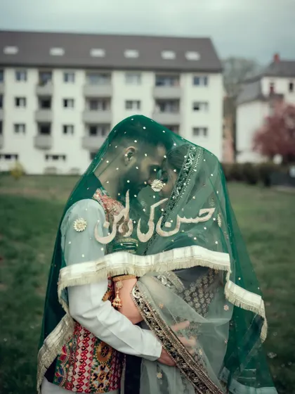 An intimate portrait of a Muslim couple under a custom green veil that reads "Hassan ki Dulhan" (Hassan's Bride).
