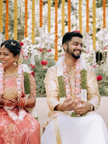 A couple shares a smile during their traditional South Indian wedding ceremony, surrounded by family and lush greenery.